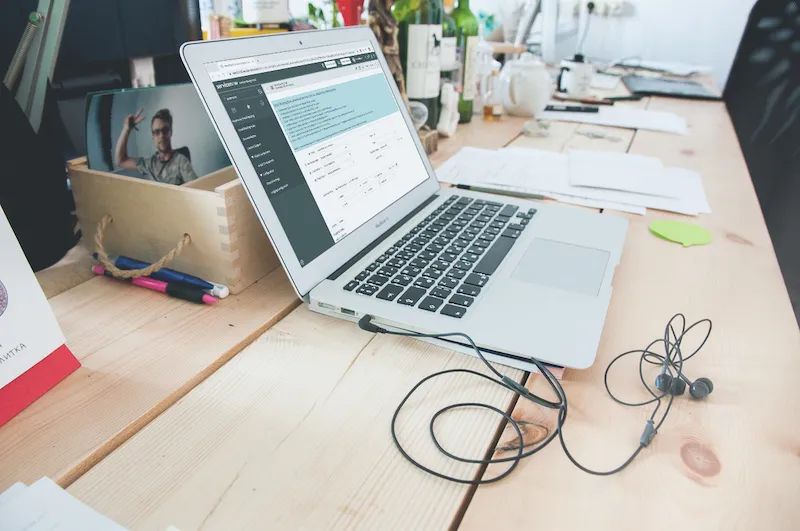 Silver MacBook laptop on a wooden desk with wired earbuds, papers, pens, and a wooden crate containing a photo of a man.